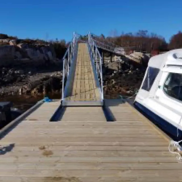 Utsikt fra brygge opp landgang med rekkverk mot land, båt til høyre.