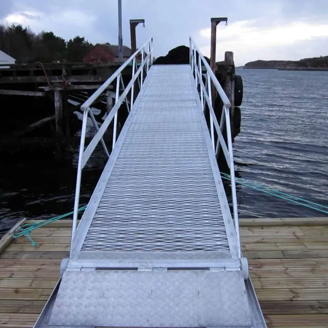 Metallgitterlandgang fører fra flytebrygge til kai ved åpen sjø.