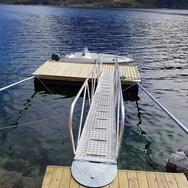 Metallrammet landgang fører til liten flytebrygge med båt i klart vann.
