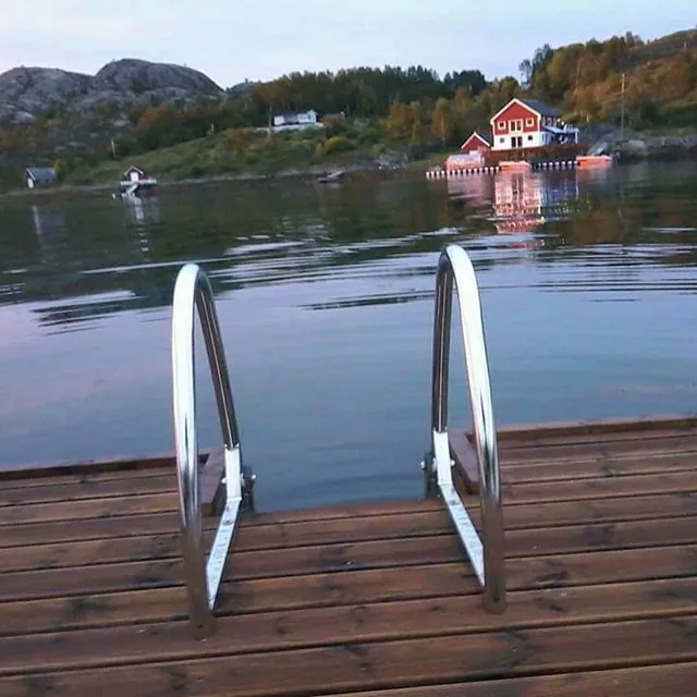 Badeplattform med metallstige ned i stille vann, rødt hus og naust i bakgrunnen.