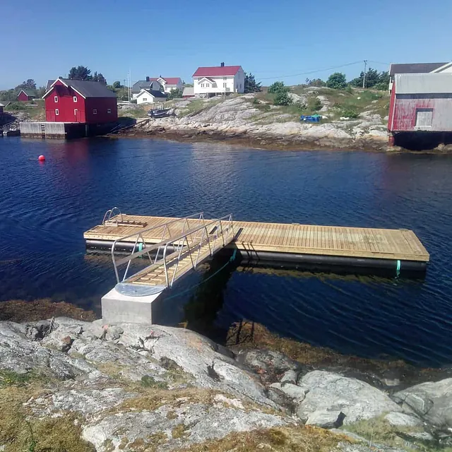 Flytebrygge med landgang fra svaberg, røde naust i bakgrunnen.