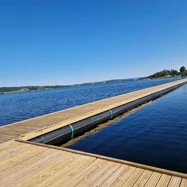Lang trebrygge med sorte flyter strekker seg ut i stille sjø.
