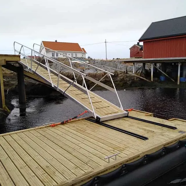 Flytebrygge med landgang opp til kai ved røde sjøhus.