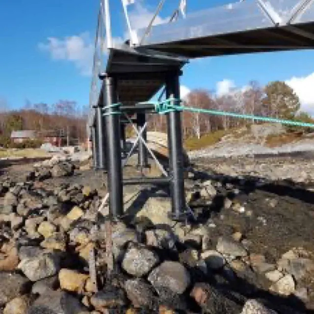 Nærbilde av landgangsfeste med sorte stolper og tau ved steinete strandkant.