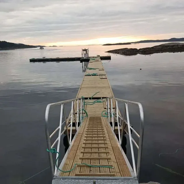 Landgang leder ut til smal trebrygge på stille sjø ved solnedgang.