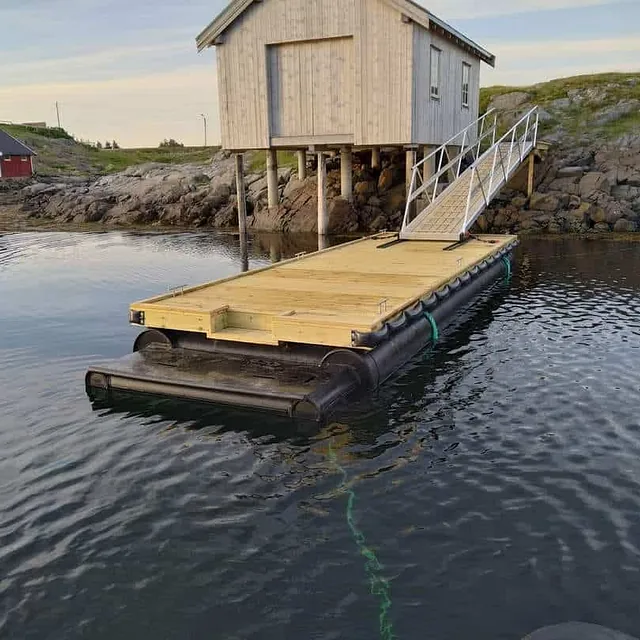 Flytebrygge forankret til trehus på påler ved steinete strand.