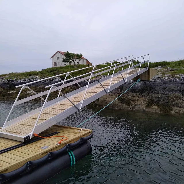 Flytebrygge med lang landgang opp til svaberg, hvitt hus i bakgrunnen.