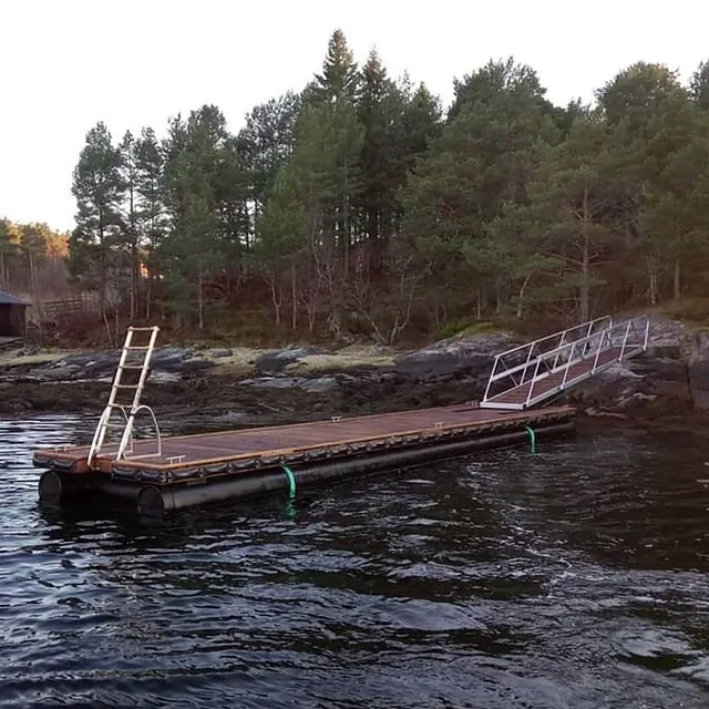 Flytebrygge med stige og landgang festet til steinete strandkant med furuskog.