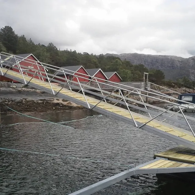 Lang metall- og trelandgang skråner ned mot flytebrygge ved røde sjøhus.